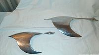 Pair of two flying geese wall sculptures in chrome finish placed on white fabric background, showing front side and size difference