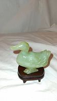 Front-side view of the smooth, light green jade duck on a dark wooden stand showing details of the carving and translucence of the jade.