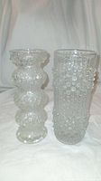 Photo of two vintage clear glass vases side by side on white fabric background. Left vase has bulbous stacked textured design, right vase is taller with vertical bubbles texture.