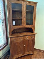 Full view of two-piece vintage oak wood display cabinet near window showing upper glass door section and lower wood door with drawers.