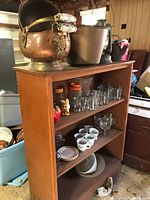Full view of wooden shelf with glassware including drinking glasses, stem glasses, mugs, small bowls, copper bucket, and copper items on top.