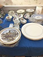Collection of various china items including floral decorated Limoges cups and saucers, stacked plates with gold trim, Wedgwood items on a blue cloth surface, and a Japanese vase in the background.