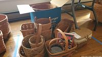 Photo showing eleven Longaberger baskets of varying sizes placed inside and around a box, some with handles visible, set near a window on the floor.