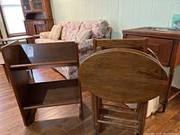 Wooden two-tier magazine rack and four TV tray tables in wooden holding rack, all placed on wooden floor, showing overall items with some furniture in background