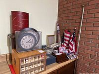 Photo showing office supplies on wooden desk against wall, including metal mesh clock, wooden organizer, red cylindrical can, American flags with pole, and multiple small office items.
