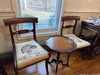 Full view of the pie crust table alongside two needlepoint upholstered wooden chairs placed by a window. Shows chair backs and needlepoint seat designs.