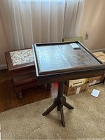 Wooden pedestal table with square raised-edge top with visible scratches next to vintage commode with floral quilted top, placed in a carpeted room near window curtains.