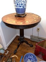 Full view of the round wooden pedestal table with a floral appliqué surface and a blue and white lamp on top.