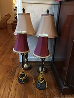Four table lamps shown in two matching pairs; two with red shades and two with beige shades, placed on floor near wall and furniture.