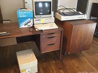 Full scene of Tandy computer system setup on wooden desk including computer tower, keyboard, CRT monitor, printer, and box of printer paper.