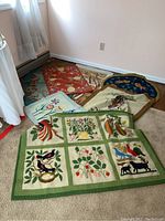 Photo showing seven rugs and tapestries arranged on the floor, including floral and bird motifs, some with green borders, others with intricate colorful designs.