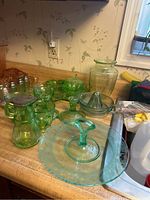 Wide view of multiple uranium glass pieces on countertop, including rectangular tray, sugar bowl, creamer, syrup dispenser, juicer, candlesticks, ring holder, shakers