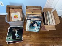 Overview photo of vinyl records boxed and stacked, showing a mix of large albums and 7-inch records in cardboard boxes and plastic tubs.