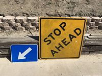 Both road signs placed side by side on ground outdoors, showing relative size and wear (yellow 'STOP AHEAD' larger with wear, blue arrow smaller and clean).
