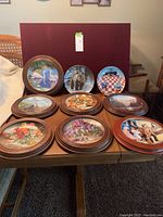 Overview of all 9 collectible plates on a table; 7 framed in dark brown wooden round frames, 2 without frames. Plates have various detailed designs including bear, swans, food, flowers.