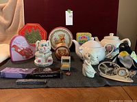 Photo showing the entire lot arranged on a surface, featuring tins, bunny bank, tea pots, clocks, figurines, and watch box.