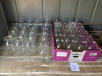 22 clear glass mason jars in groups, half-pint size, set on wooden surface, including jars in cardboard box and loose ones.