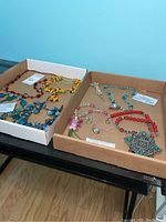 Overview of seven colorful necklaces displayed in two cardboard boxes on a table, showing variety in materials and styles