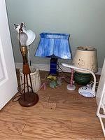 Five desk lamps placed together on wooden floor showing various sizes, designs, and materials with a small white hand towel rack on the side.