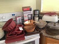 Countertop with various kitchen items including Black and Decker single cup coffee makers in box and out, silicone baking molds, wicker basket, baking trays, pitcher, and stainless steel cups.