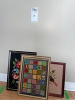 Three framed embroidered artworks leaning against a wall including a colorful patchwork needlework, a black floral embroidery, and a third framed embroidery with floral and bird design.