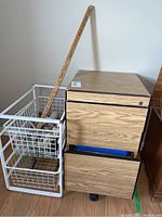 View showing small wood laminate file cabinet and adjacent white wire basket storage rack with three baskets, wooden yardstick leaning against them.