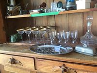 Shelf photo showing 12 gold-rimmed stemmed wine glasses, 8 molded glass goblets, crystal decanter, stainless steel meat platter, butter dish, and some mugs.