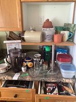 Photo of shelf with various kitchen items including containers, pitchers, mugs, a kitchen appliance attachment, and bowls.