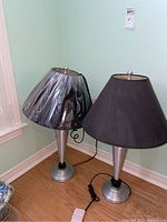 Pair of two matching table lamps with silver metal conical bases and black fabric shades. One shade has plastic wrap covering.