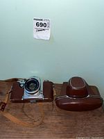 Camera body with brown leather half-case and separate top-cover case on wooden surface beneath lot tag