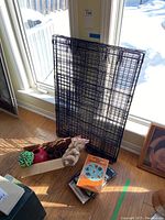 Folded wire pet crate with pet toys, leashes, collars, and books arranged on the floor near a window.