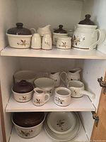 Full set arranged inside white cabinet shelf showing upper and middle shelves with various pitchers, covered casserole, teapot, salt and pepper shakers, and bowls