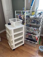 Wide view showing two metal wire storage units filled with various household items and a white plastic drawer unit with a laundry basket on top