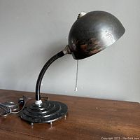 Side view showing the full desk lamp with flexible gooseneck, stepped circular base on four feet, and black rounded globe shade with visible wear and marks.