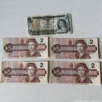 Photo showing one Canadian 1-dollar bill at top and four Canadian 2-dollar bills below on white background.