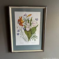 Framed print showing colorful wild flowers with a silver frame and blue-grey mat against a grey wall.