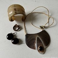 All four jewelry pieces shown together on white surface including the carved cuff bracelet, wooden ring, abalone pendant with cord, and seed earrings.