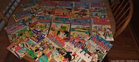 Full view of 20 vintage comic books spread out on a round wooden dining table showing colorful covers with characters from Archie, Betty Veronica, Josie, and others. Various issues visible with bright colors and classic mid-20th century comic styling.