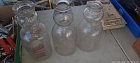 Three clear glass antique milk bottles with baby face design, different label styles sit on a table.