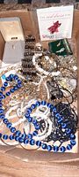 Top view of the lot showing various gold-tone jewelry pieces, a jewelry box with a pendant, white and blue strand necklaces, and the American Heart Association red dress pin.