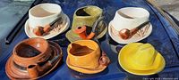 Six ceramic hat containers with attached pipes in varied colors and styles, displayed on a dark surface