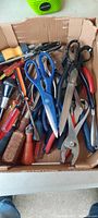 Box containing various hand tools including scissors, pliers, wire cutters, and screwdrivers with mixed colored handles.