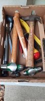 Top-view photo of a box containing multiple carpenter hand tools including two wooden handled hammers, wire brushes, and other various tools.