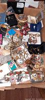 Overview of box filled with assorted vintage and modern costume jewelry, including rings, bracelets, earrings, cameos, and various pieces, showing diversity and mix of colors and materials.