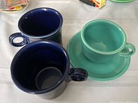 Top view of two dark blue mugs and one turquoise cup with saucer, showing size and color differences.