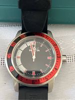 Front view of Invicta men's watch face with black dial, red and black bezel, white and red hour markers and hands, and black rubber strap attached.