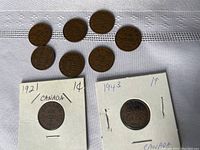 Eleven small Canadian pennies arranged on a fabric surface, two coins individually placed in holder sleeves showing dates 1921 and 1943. Other coins laid out above these holders show reverse side with maple leaves and
