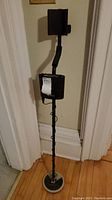 Full view of the metal detector showing the black shaft with armrest and circular search coil at the bottom standing on a wooden floor against a wall.