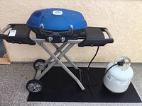 Full view of portable Napoleon grill with blue lid, foldable legs, side shelves, attached propane tank and protective mat beneath