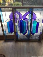 Two decorative stained glass panels leaning against a window, showing abstract purple and blue designs in wood frames.